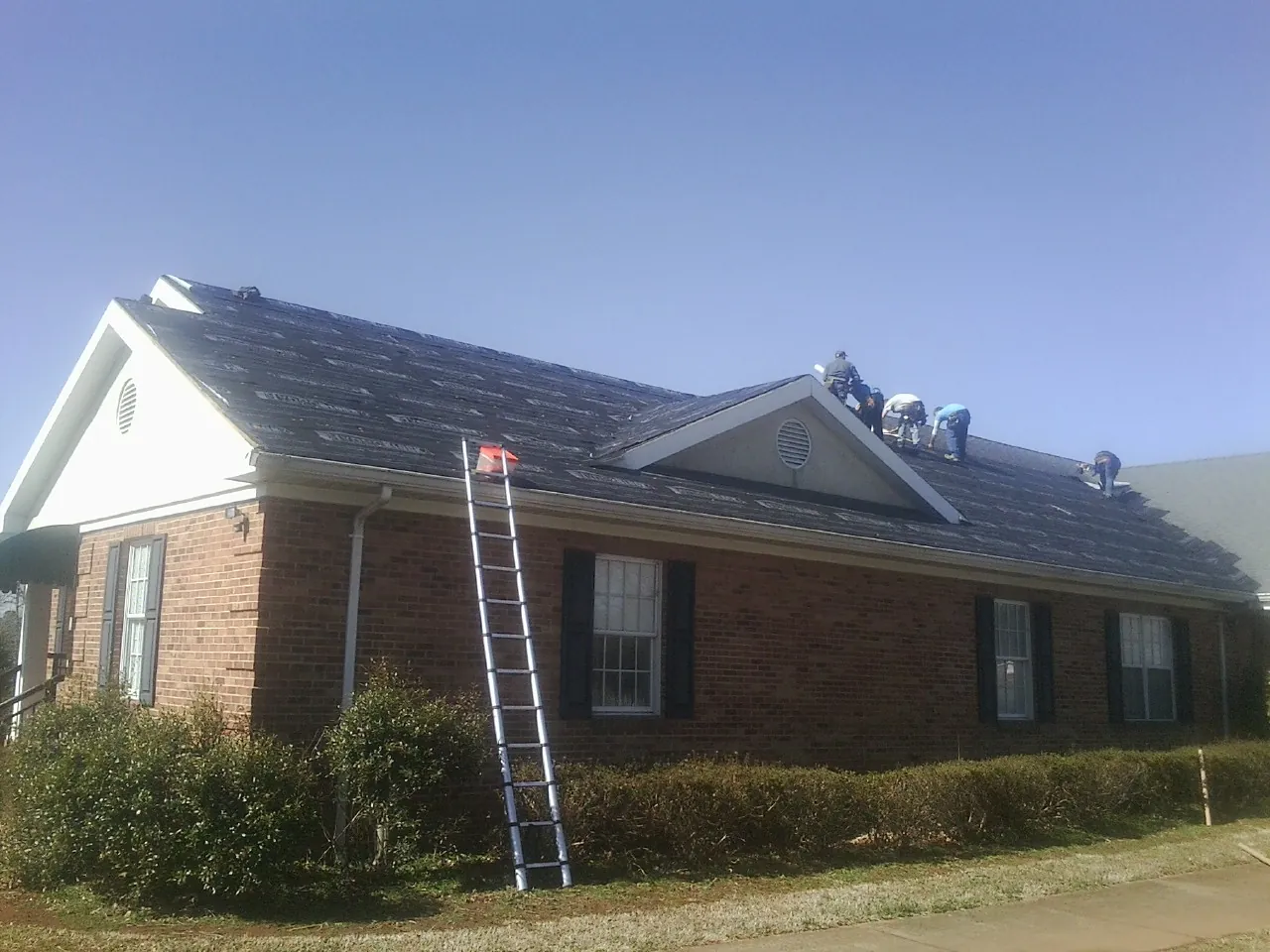 Crew tearing shingles off a brick ranch roof with a ladder set — Metal Roof Repair work by our crew in Radford