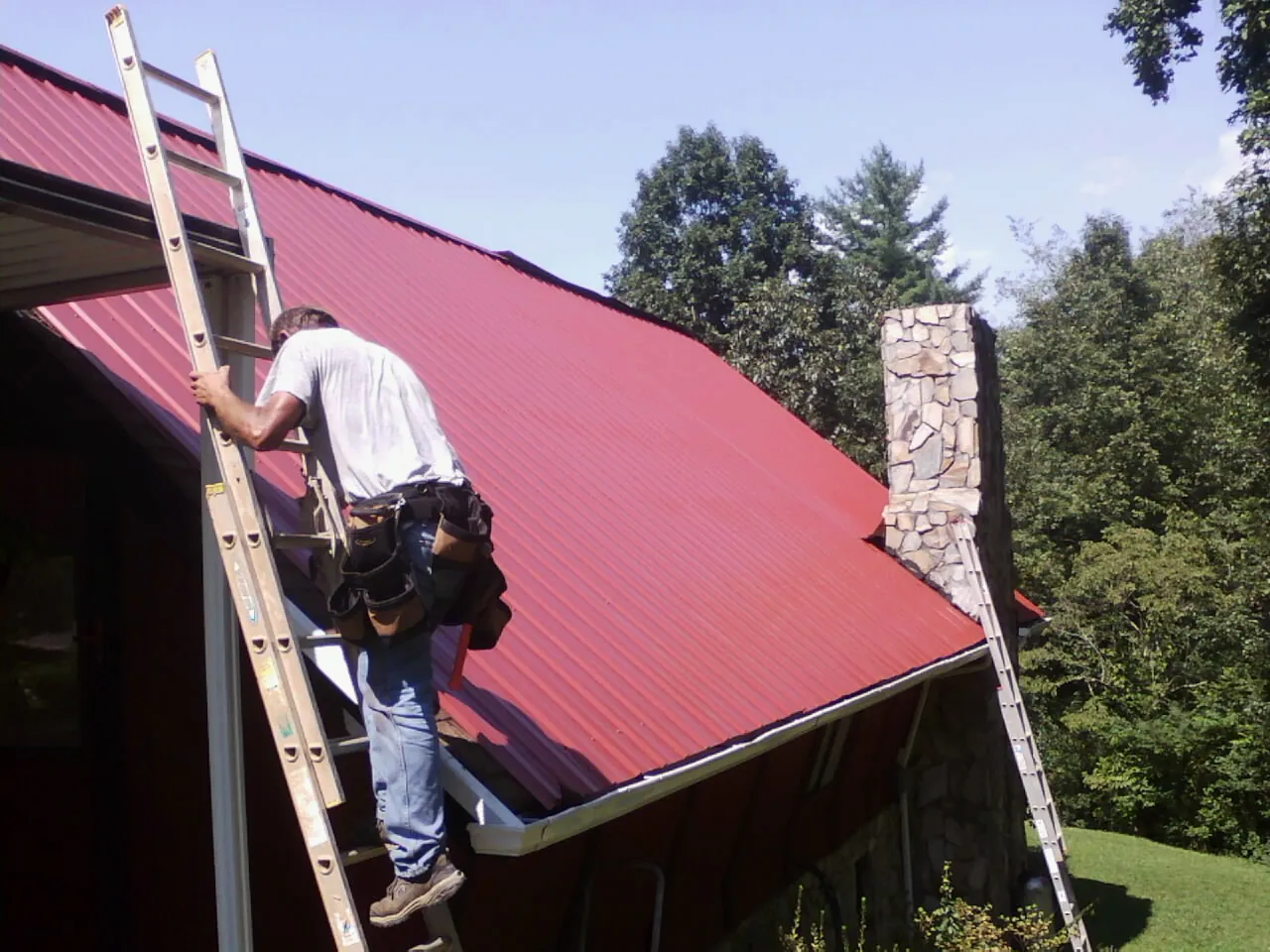 Roofer on a ladder beside a red metal roof and stone chimney — Soffit & Fascia Repair by Roofing Brothers in Radford